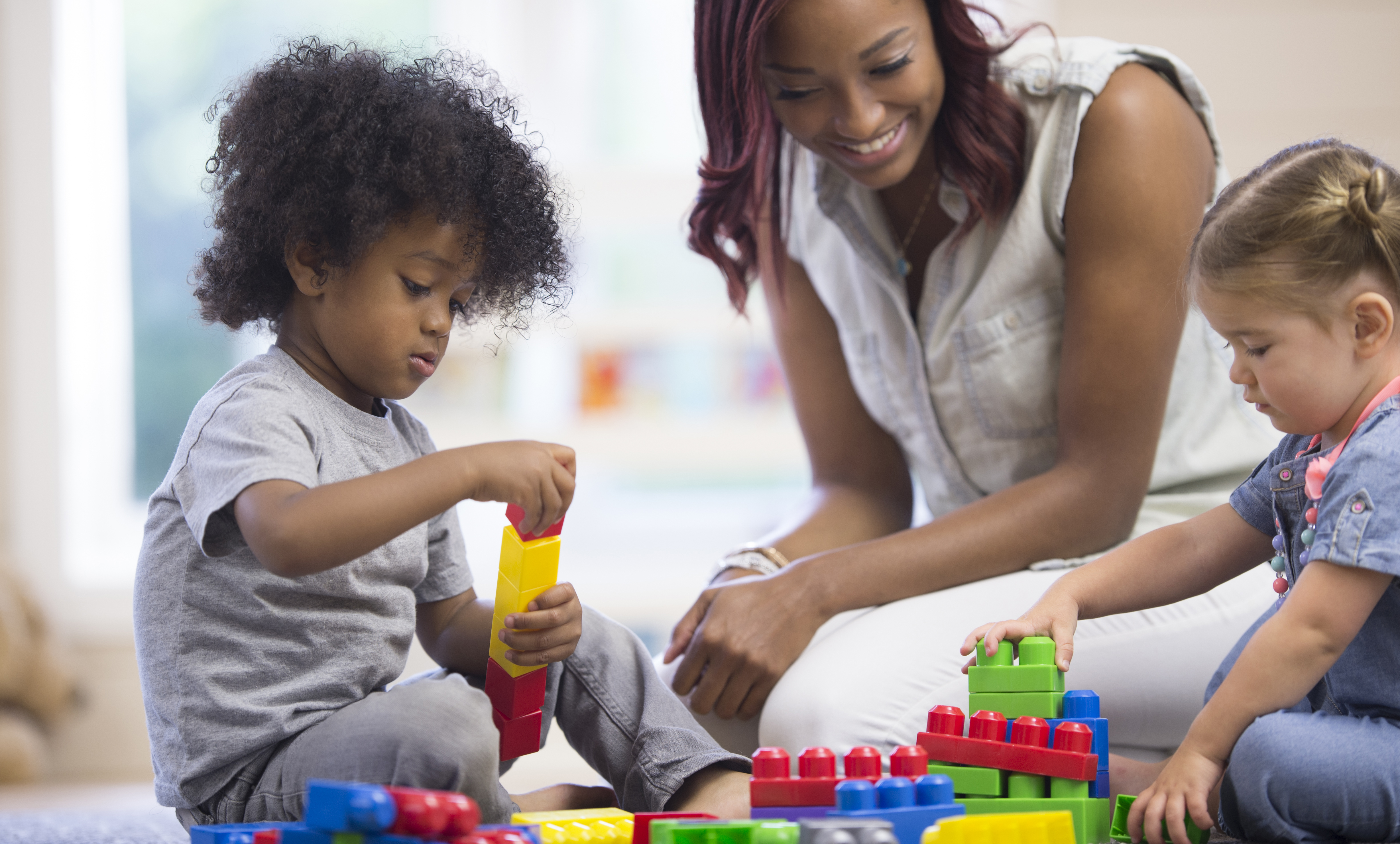 A teacher is sitting on the floor with her preschoolers and day care students - they are playing with plastic blocks together.