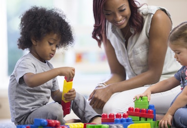 A teacher is sitting on the floor with her preschoolers and day care students - they are playing with plastic blocks together.