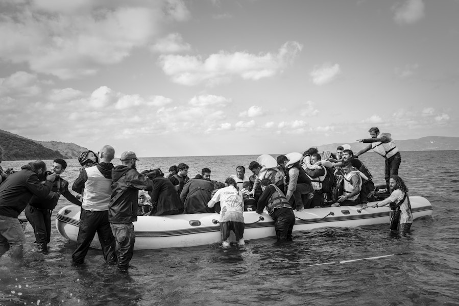 Lesbos, Greece - October 25, 2015: Volunteer lifeguards and others assist migrants out of their boat after landing on the Greek island of Lesbos, near the town of Skala Sikamineas. A photographer (left) photgraphs the landing. The coastline of Turkey is visible on the rightside of the photo.