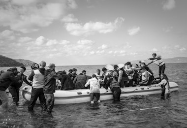 Lesbos, Greece - October 25, 2015: Volunteer lifeguards and others assist migrants out of their boat after landing on the Greek island of Lesbos, near the town of Skala Sikamineas. A photographer (left) photgraphs the landing. The coastline of Turkey is visible on the rightside of the photo.