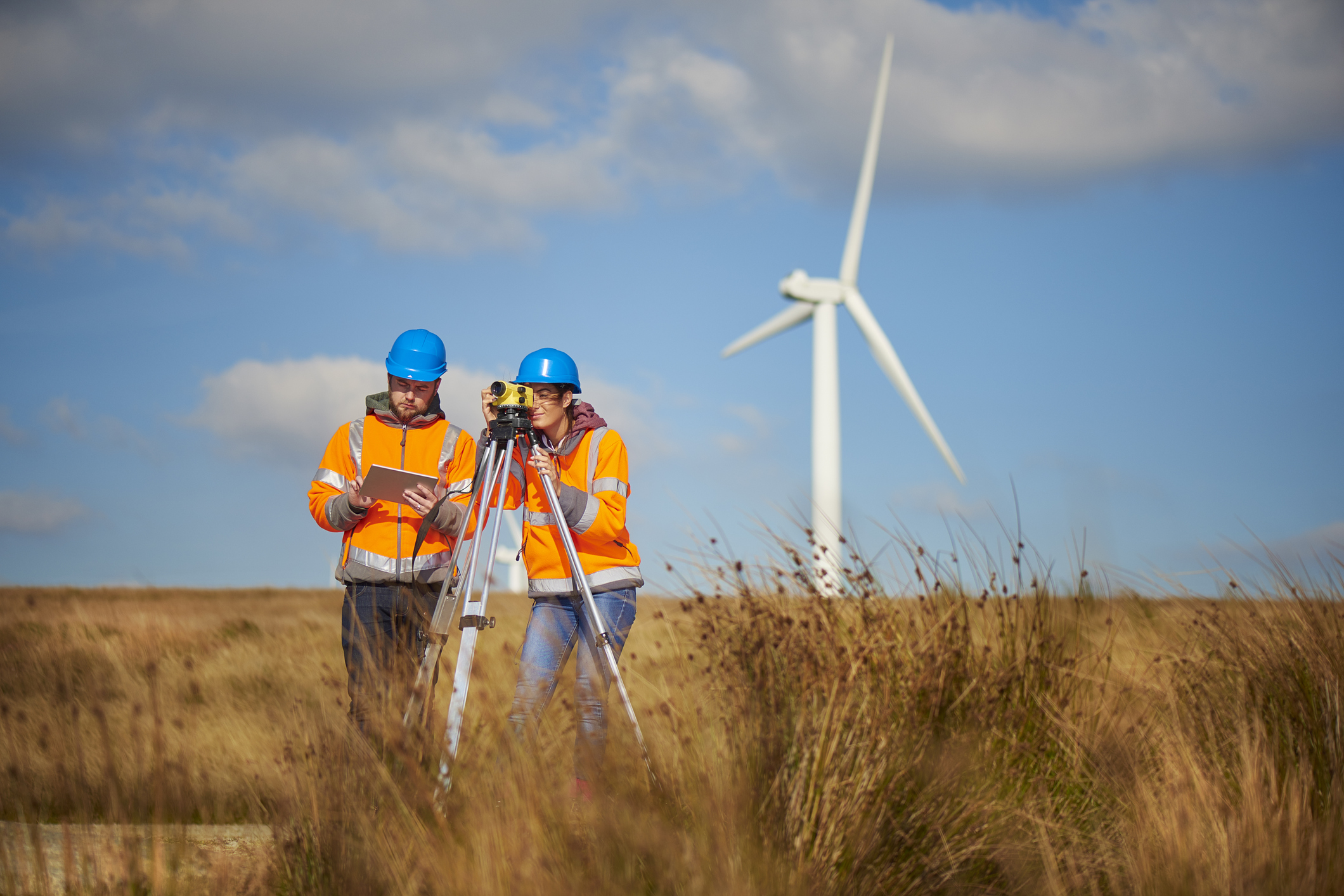 two wind farm engineers using a builder's level to plan out the expansion of the wind farm site. they are wearing orange hi vis jackets and blue hard hats . one is male , one is female. In the background wind turbines can be seen across the landscape.
