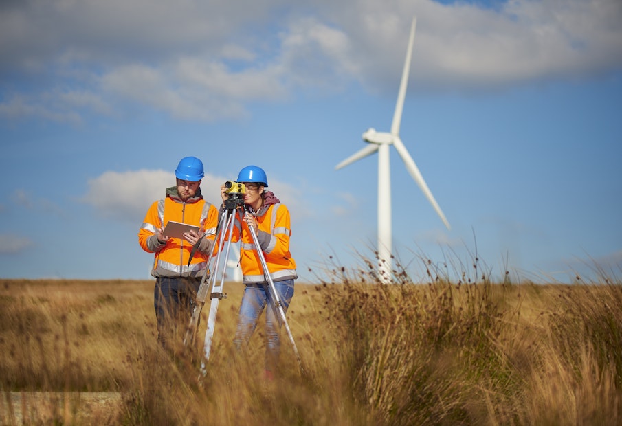 two wind farm engineers using a builder's level to plan out the expansion of the wind farm site. they are wearing orange hi vis jackets and blue hard hats . one is male , one is female. In the background wind turbines can be seen across the landscape.