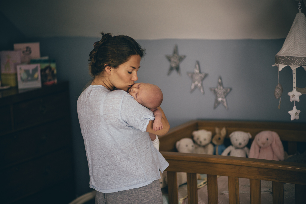 A happy new mother kisses her newborn baby on the cheek as she gets ready to put her down in the cot to sleep. The bedroom has stars on the walls and there are soft toys in the cot.