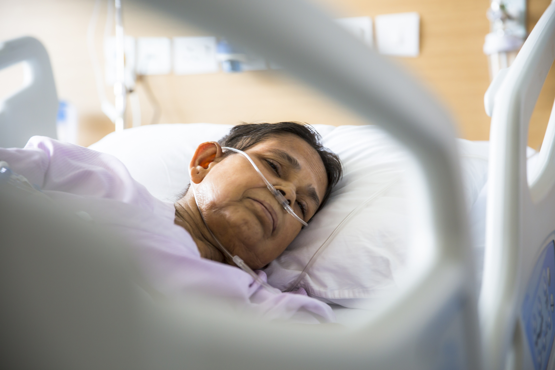 Old woman patient lying on Hospital bed with Oxygen tubes in her nose. She has her eyes closed.