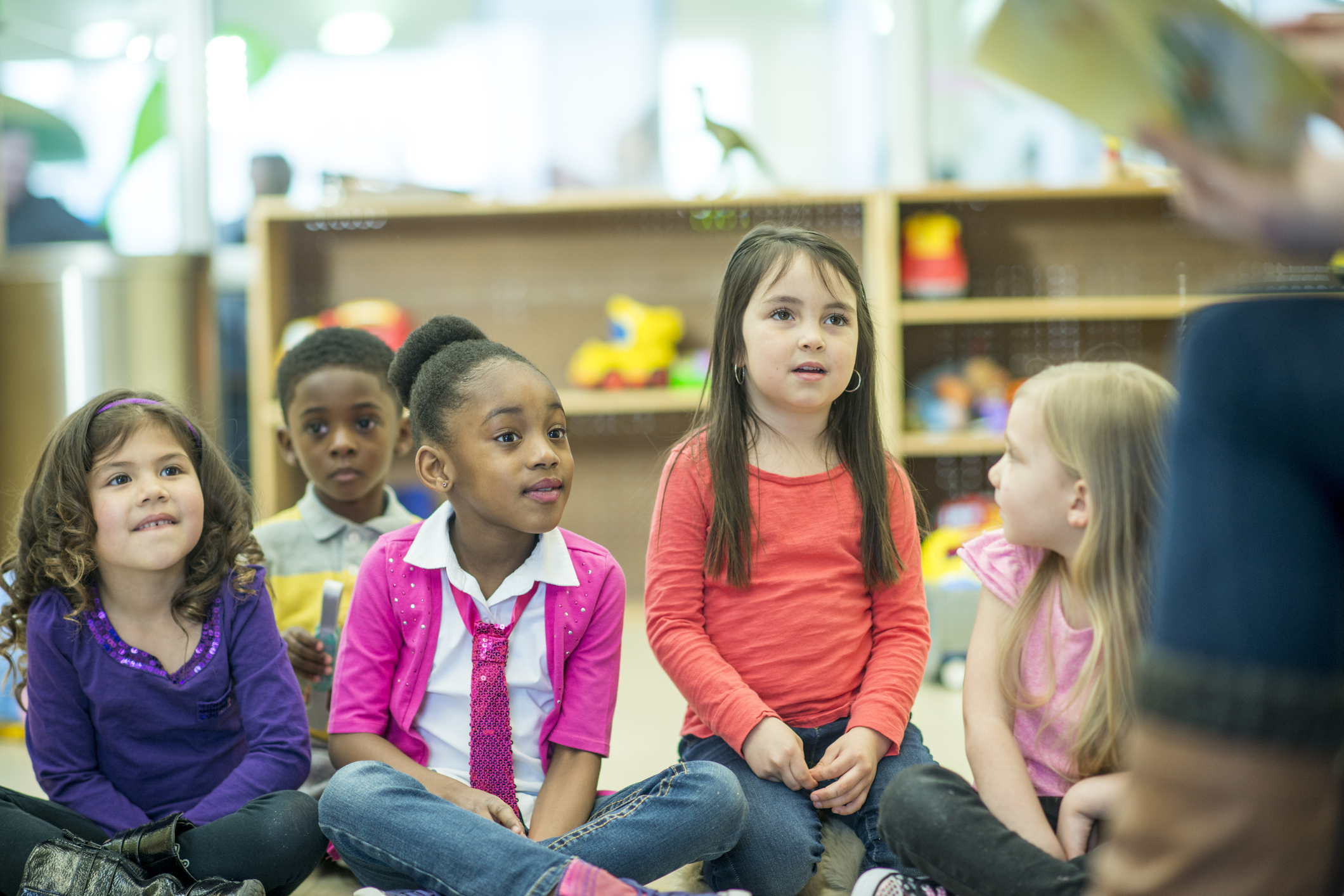 A multi-ethnic group of elementary age children are listening to their teacher read them a book in the library.