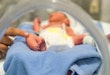 Photo of a premature baby in incubator. Focus is on his feet and toes. The doctor is touching him to check his reflexes. There are cables and tubes in the out-of-focus area.