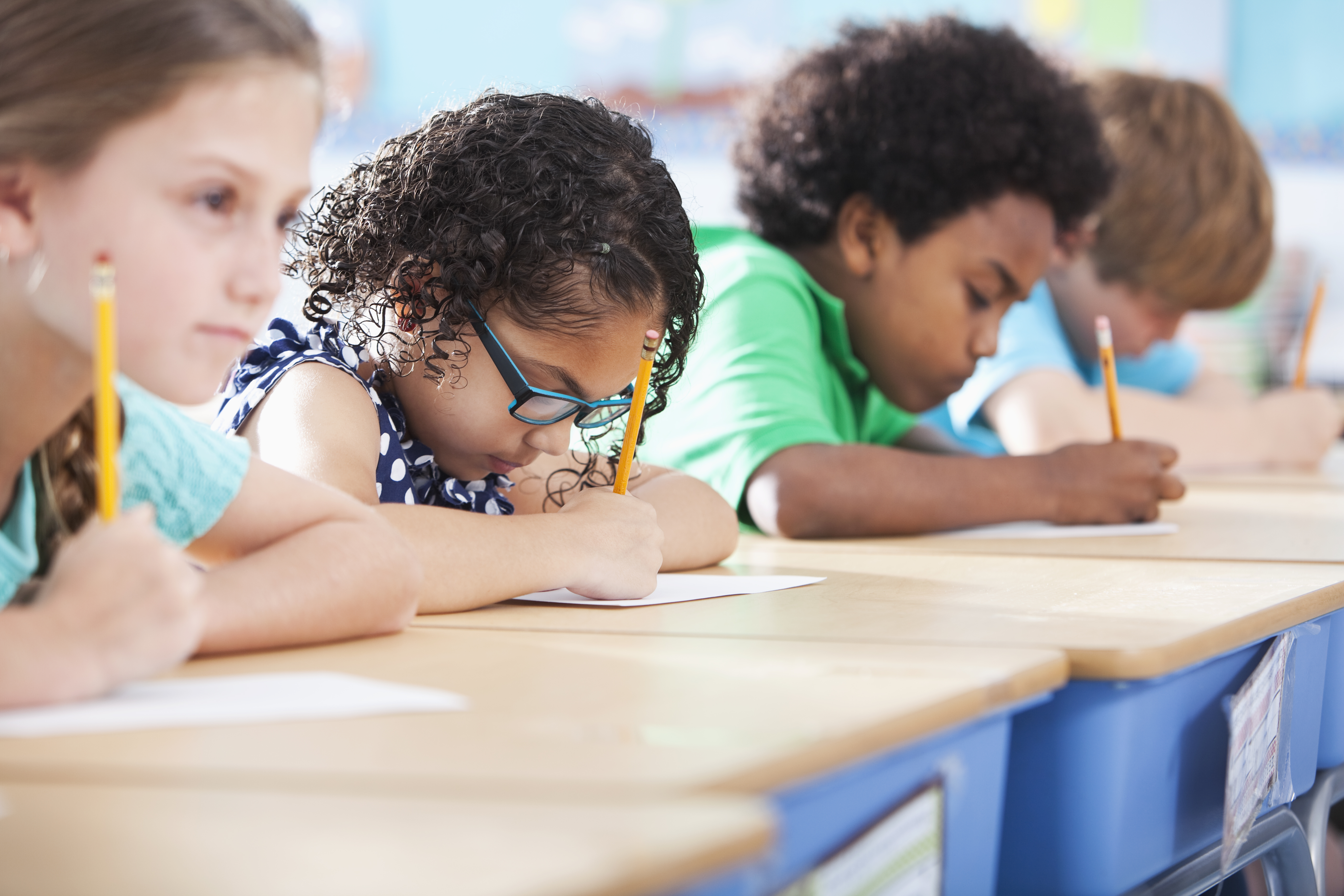 Multi-ethnic elementary school children writing in classroom.  Focus on Hispanic girl wearing eyeglasses (8-9 years).