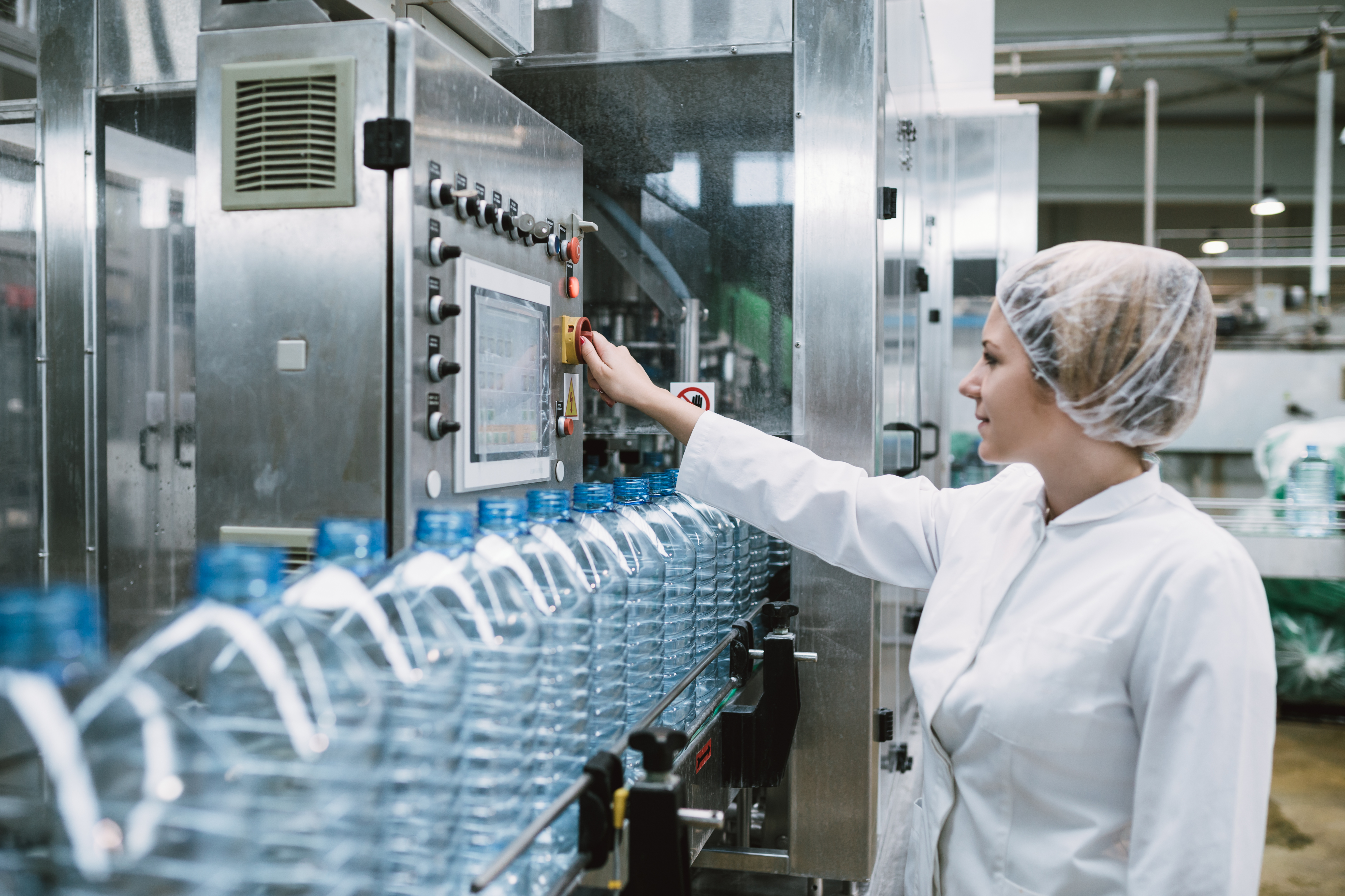 Young happy woman worker checking robotic line for bottling and packaging pure drinking water into bottles and canisters.
