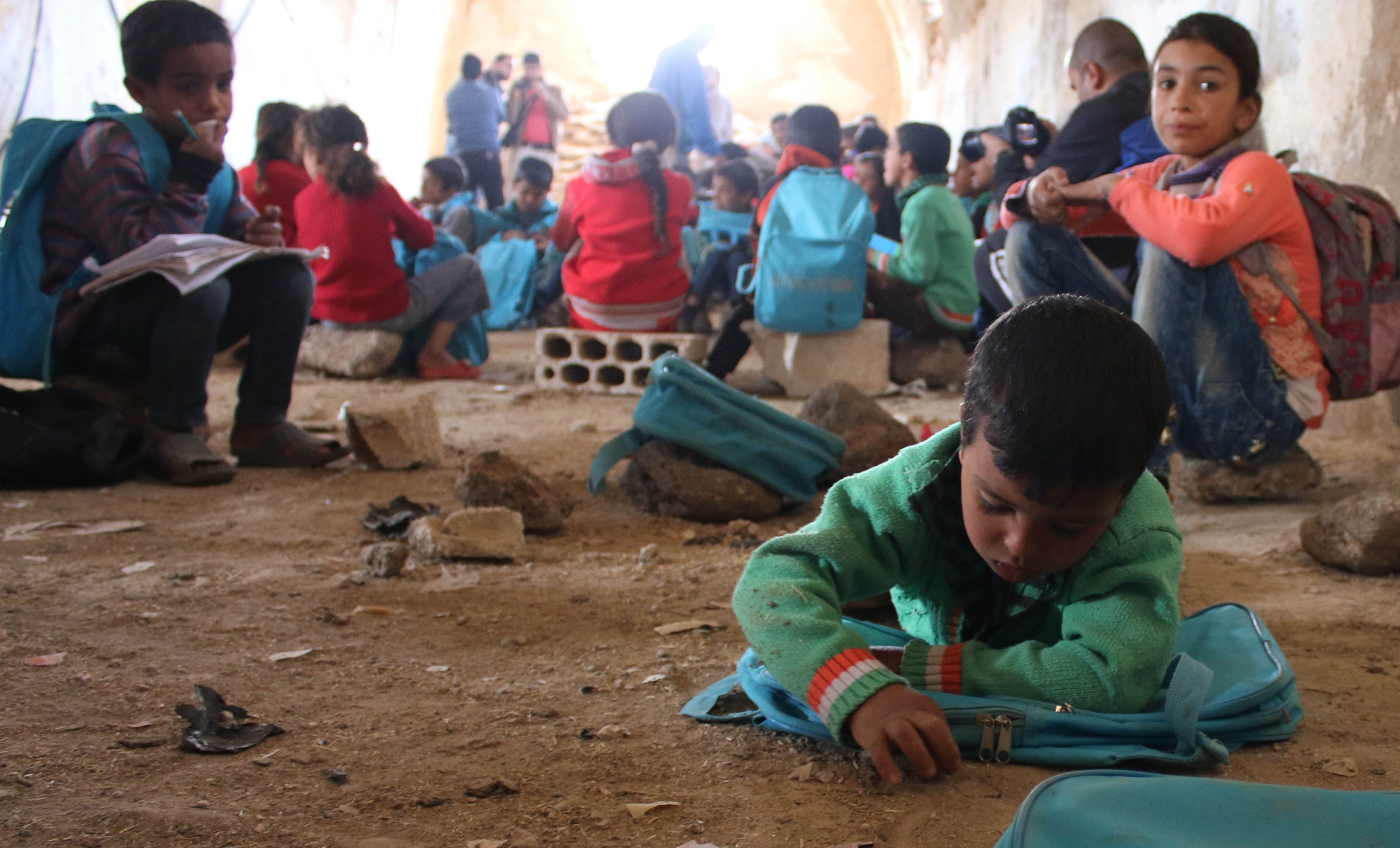 On 13 November 2016, children lie or sit on the ground writing in notebooks at a make-shift school in rural Dar'a in the Syrian Arab Republic. Despite the ongoing violence across the country, children and dedicated teachers are doing all they can to keep their education going. Some 1.7 million children are currently (Nov 2016) out of school in the Syrian Arab Republic. Schools across the Syrian Arab Republic continue to be destroyed and damaged. This year (2016) 84 attacks on schools have resulted in the deaths of 69 children. UNICEF has provided 3.2 million children with learning materials, such as school bags, stationary and text books.

In November 2016, there is no safe place for children to learn or play in Syria. Underground facilities including basements and even caves, are used to shelter children from a war they grew up knowing nothing else. Displaced families from different areas in rural Damascus and Dar’a sought refuge in tents in rural Dar’a. The make-shift school receives up to 80 children on a daily basis. They are grouped according to age and knowledge in four groups, where each of the four former teachers, displaced themselves, teaches them reading and writing in both Arabic and English and the principals of mathematics. “There is not a single space or even an extra tent that we could’ve used as a learning space,” says [NAME CHANGED] Muhammad, a teacher in this school. “So we had to clean this fodder collection centre and turn it to a school where almost 80 children come on a daily basis to learn reading and writing, both in Arabic and English, and the principals of mathematics,” Muhammad continues.  The children are divided into four groups based on their knowledge and age. Each group is taught by one of the four former teachers, two male and two female teachers. “Children have to share notebooks because we severely lack the necessary education supplies. They rotate on using the only six desks available.”  [NAME CHANGED] Mona, a