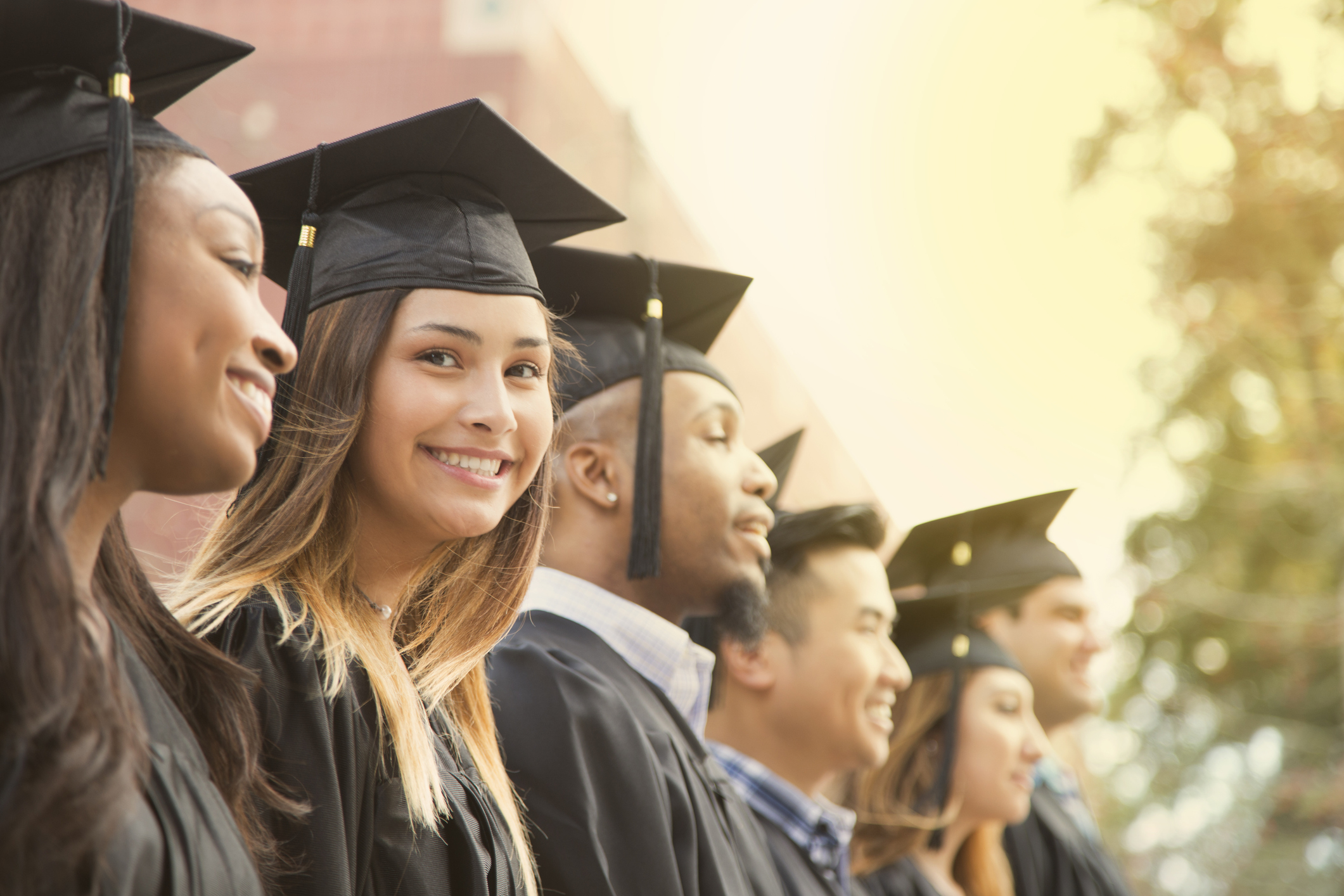Latin descent girl waits in line during college graduation ceremony.   She looks at camera with a big smile as she wears a black cap and gown.