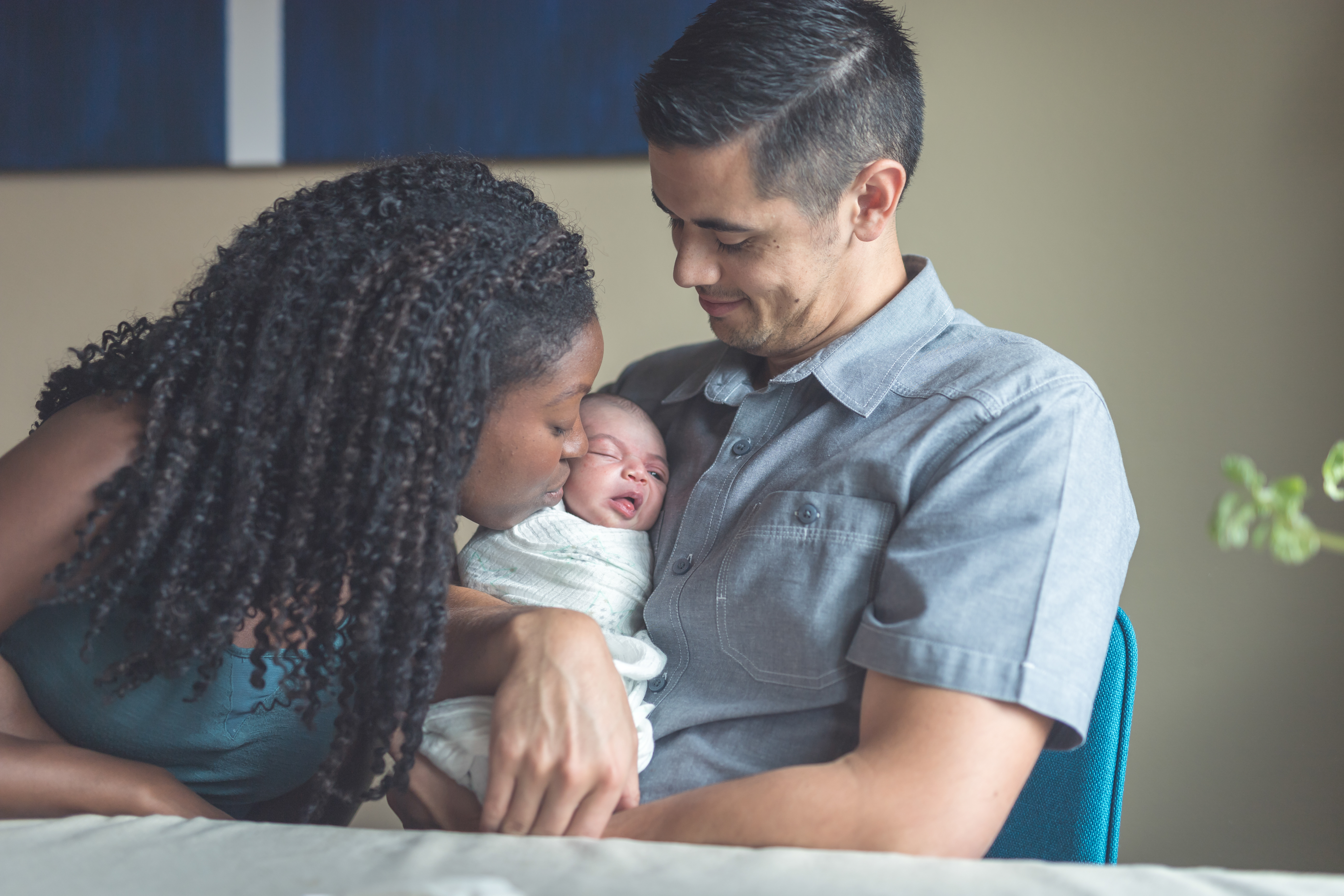 Dad holds their newborn baby girl gently as mom leans over to softly kiss her cheek. Baby is swaddled and has one eye open as she smiles. Or grimaces.
