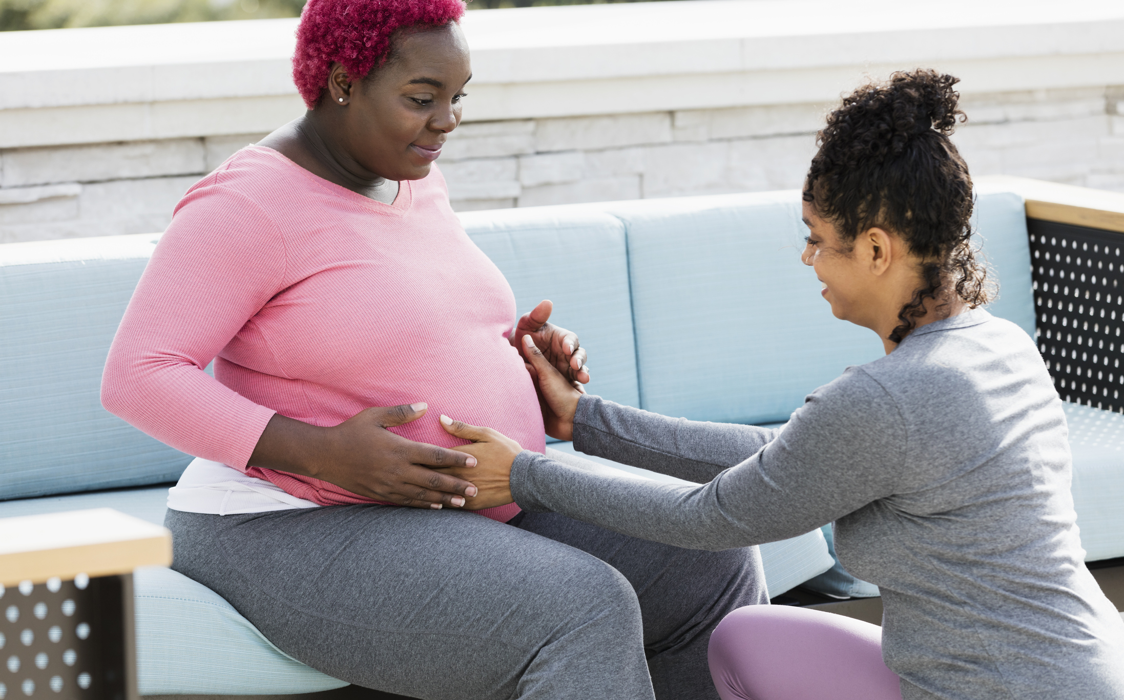 A pregnant African-American woman with her doula or birth support coach. The expectant mother, who had pink hair and is wearing a pink shirt, is sitting outdoors on a patio sofa smiling. The doula, a mixed race woman, is kneeling in front of her, touching her round abdomen. Both women are in their 30s.