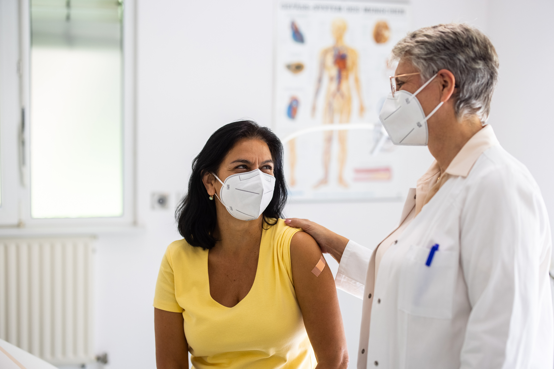 Mature woman visiting clinic for health checkup with a female doctor standing by. Senior doctor talking with female patient in clinic. Both wearing face masks.
