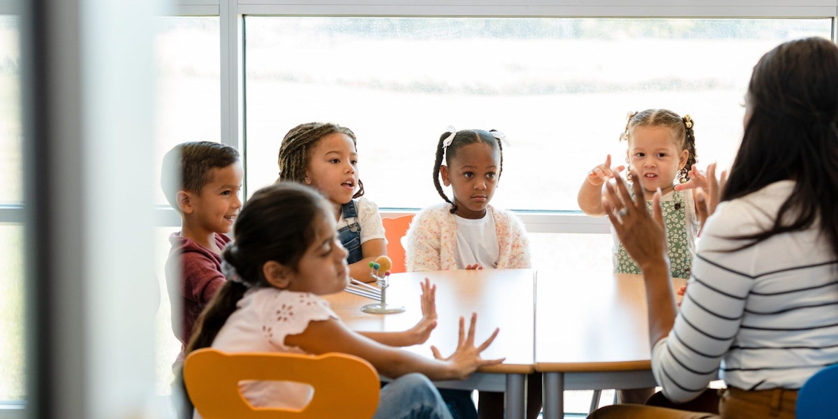 Sitting at a table in their classroom, a multi-ethnic group of preschool age children plays counting games with their unrecognizable female teacher.