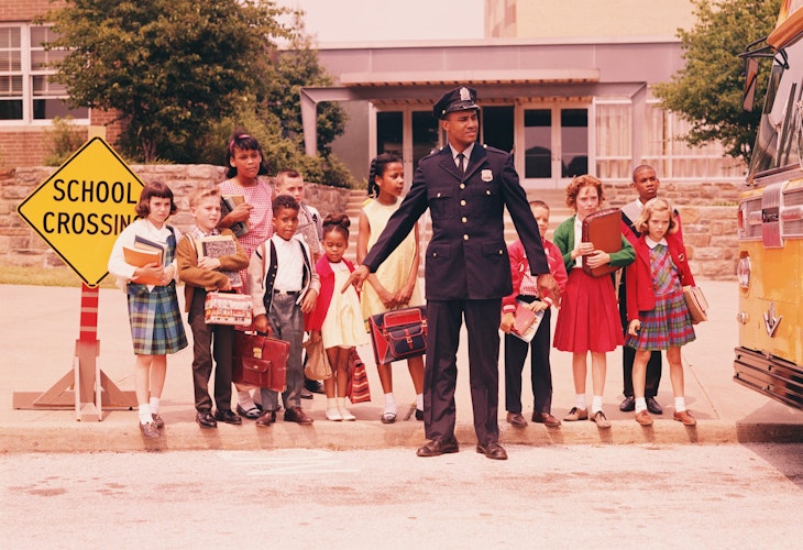 UNITED STATES - CIRCA 1960s: Group of children at curb in front of school, waiting for signal from policeman.
