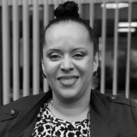 Light-complected Black woman shown shoulders up wearing hair in bun atop her head, blue button earrings, makeup with red lipstick and smiling. She is wearing olive-colored blazer and blue and white patterned blouse with long necklace of various blue-colored pendants.