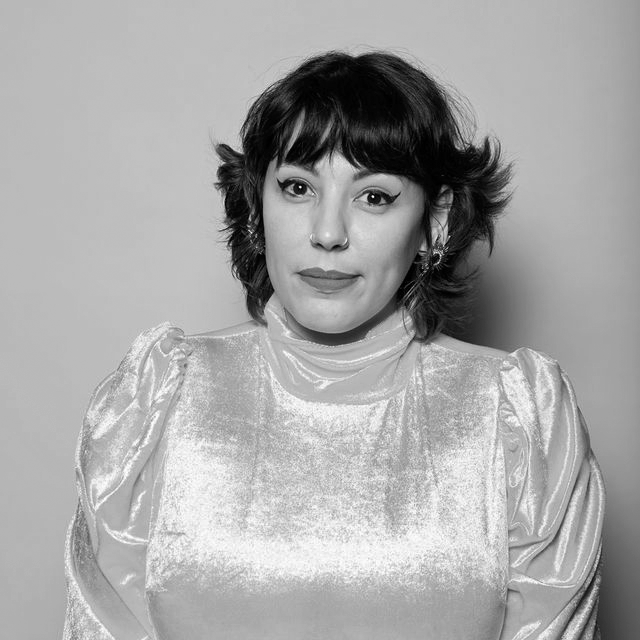 A Chicana person with short brown hair wearing bright red lipstick and a bright gold blouse.