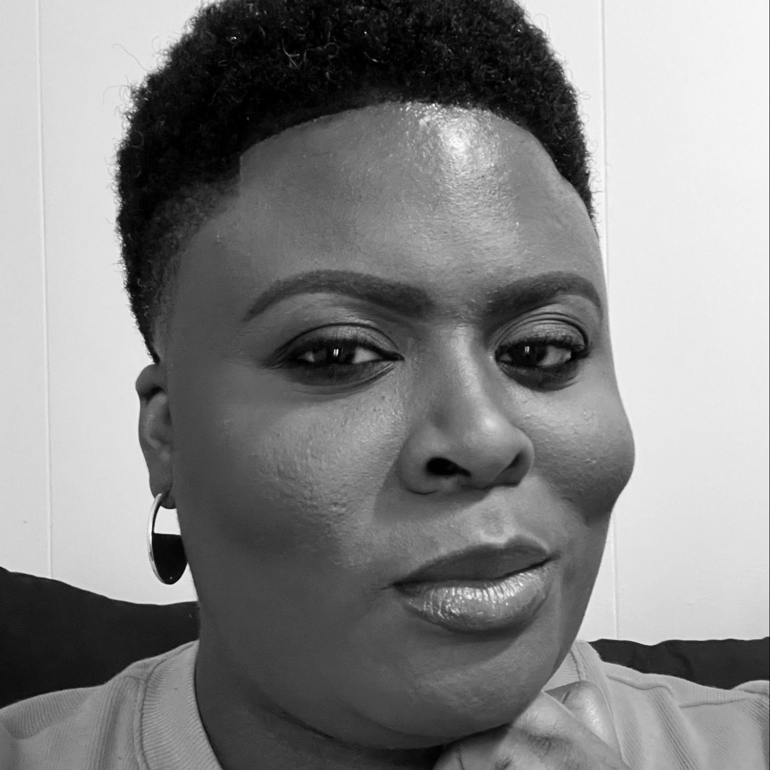 Chimére L. Smith is a Black woman with a shade faded haircut, close on the sides, about an inch on the top. She is wearing a coral short-sleeved blouse with hoops earrings and has her left hand under her chin.