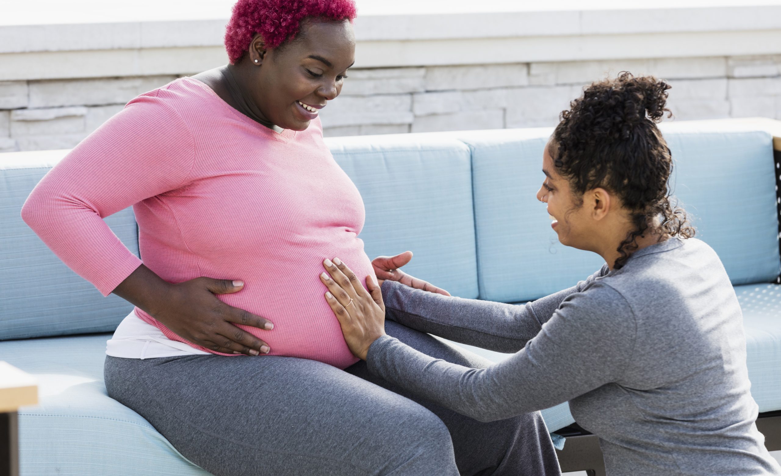 A pregnant African-American woman with her doula or birth support coach. The expectant mother, who had pink hair and is wearing a pink shirt, is sitting outdoors on a patio sofa smiling. The doula, a mixed race woman, is kneeling in front of her, touching her round abdomen. Both women are in their 30s.
