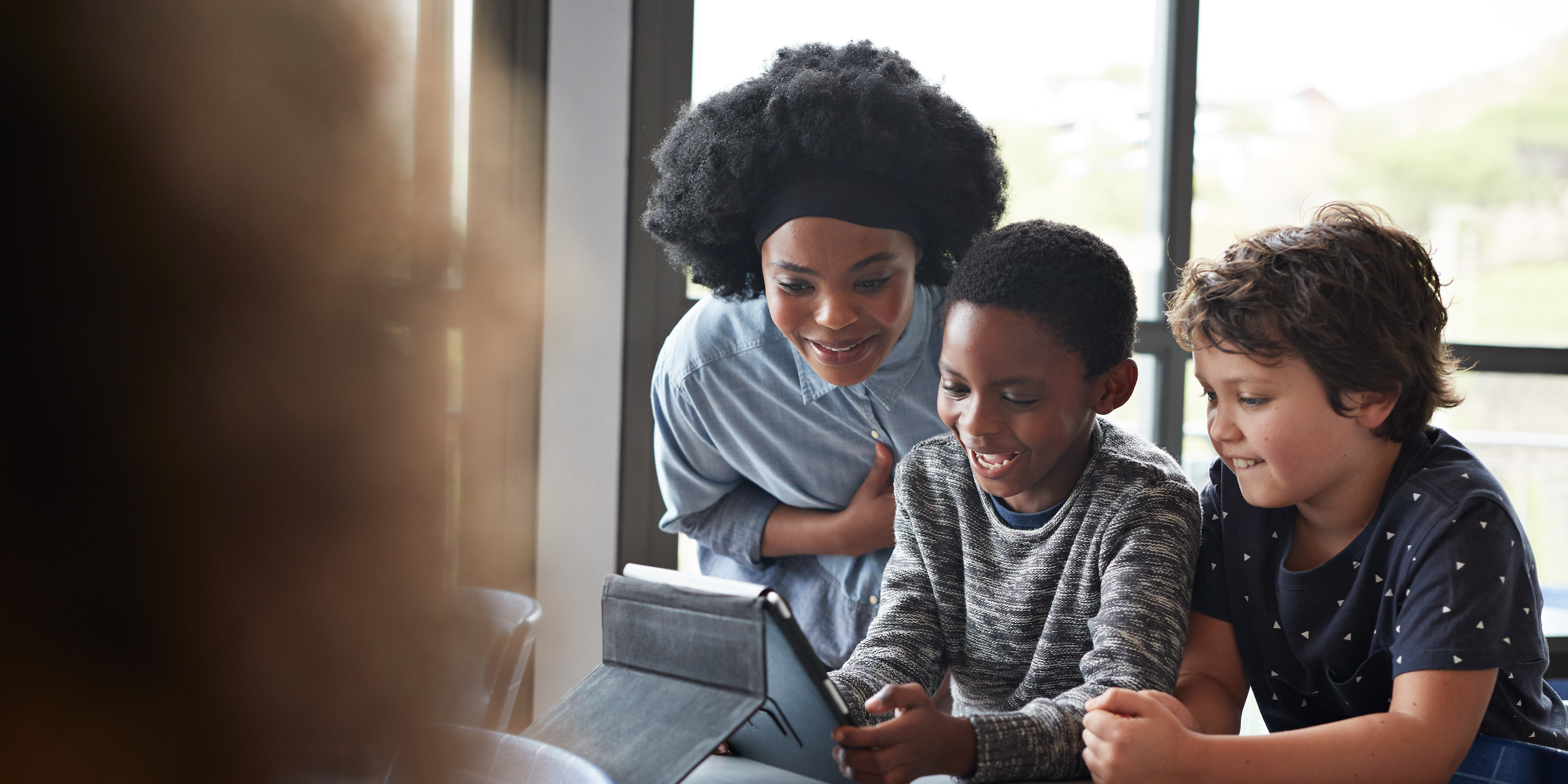 A Black teacher is looking at an iPad with two students.