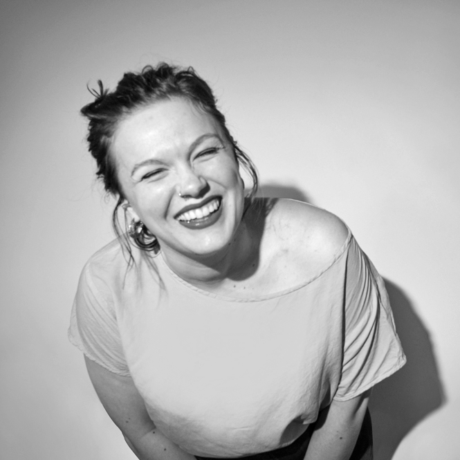 Headshot of dark-haired woman with light skin in a yellow off-shoulder top, red lipstick, and a nose ring bending over to laugh at the camera.