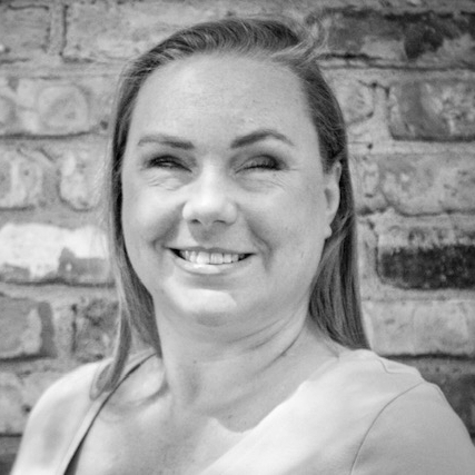 Bridgit stands in front of a brick wall, smiling. She wears a pale pink t-shirt with a deep v-neck. Her long, straight brown hair is parted on her left, and tucked behind her left ear. Her face is sunkissed and lightly freckled, and her left eyebrow is arched up as she faces the camera.