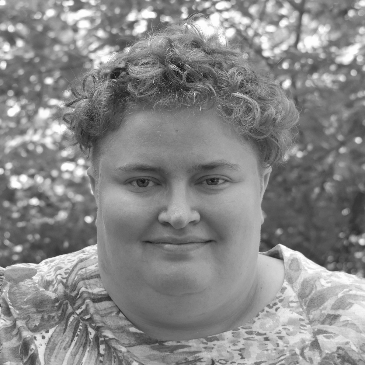 A headshot of a white person with short curly golden brown hair and blue eyes. They are wearing a leafy floral shirt. The backdrop is green summer foliage dappled with sunlight.