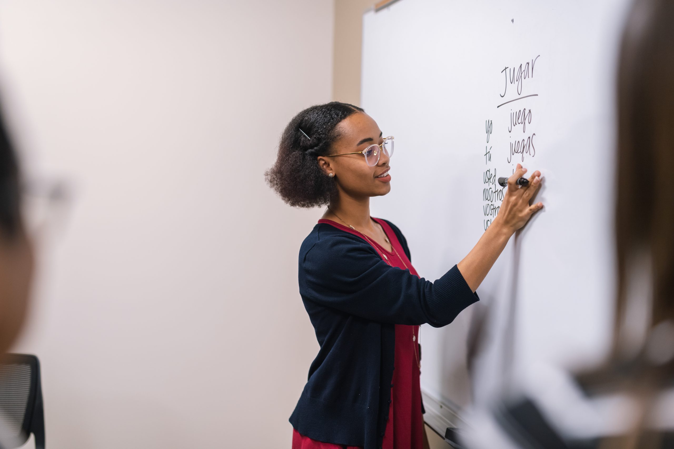 A young ethnic female professor stands at a large whiteboard and writes down Spanish verbs to conjugate.