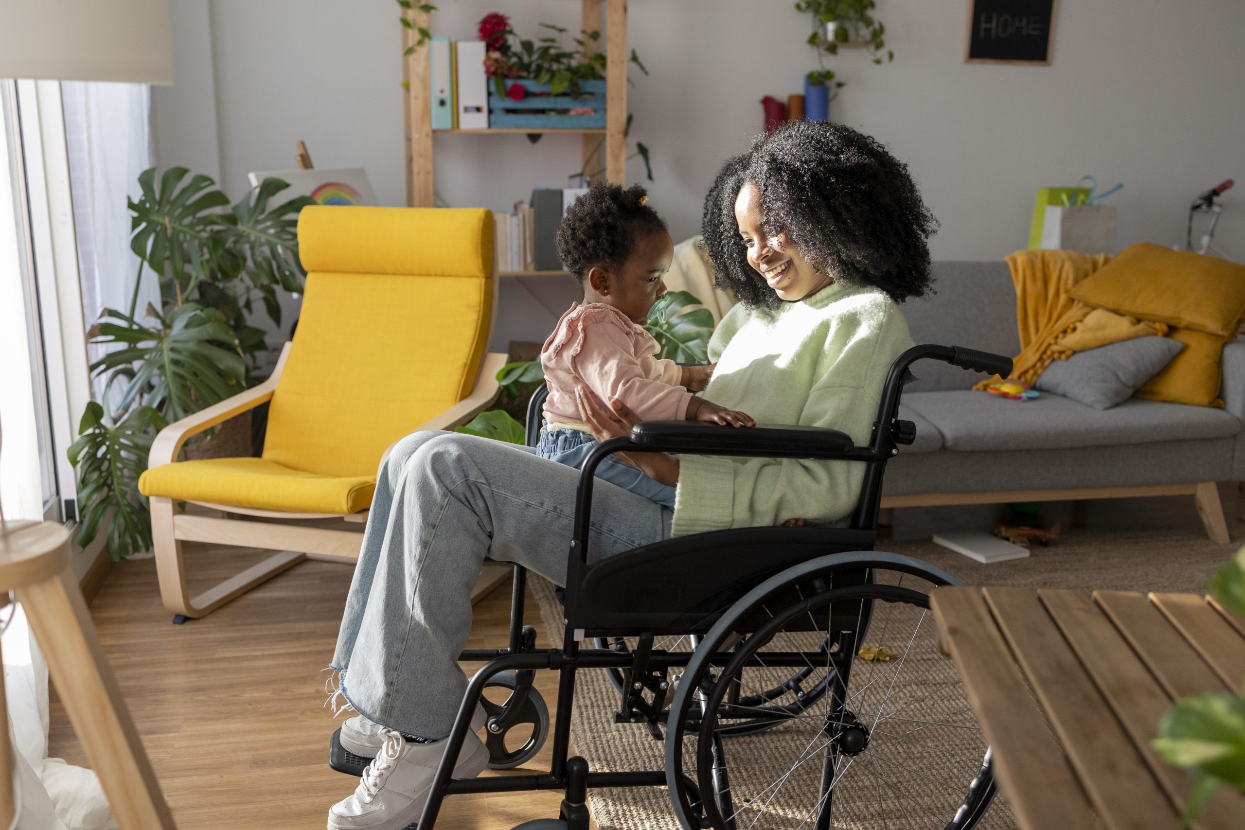 Toddler girl sitting on smiling mother with disability in wheelchair at home