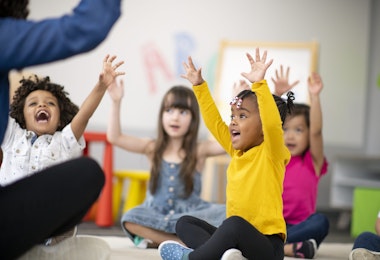 A multi-ethnic group of preschool students is sitting with their legs crossed on the floor in their classroom. The mixed-race female teacher is sitting on the floor facing the children. The happy kids are smiling and following the teacher's instructions. They have their arms raised in the air.