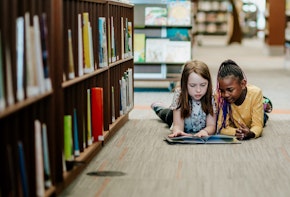 Two young girls reading a book on the floor of library