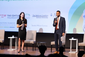 Dr. Stefan Lallinger holds a microphone on stage in front of a large projection screen.