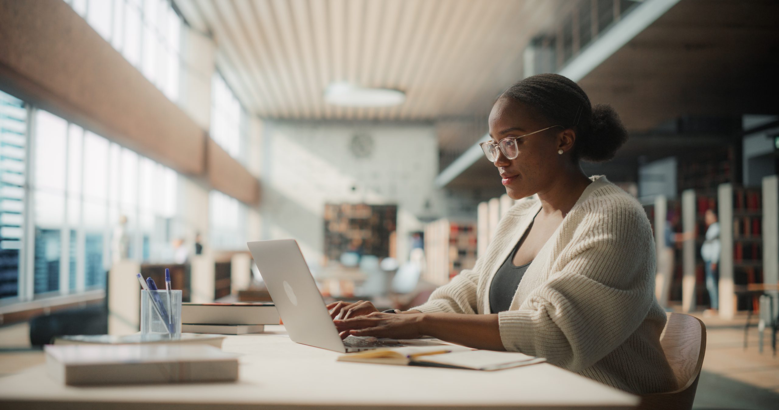 Dedicated African American Female Student Engaged in Online Learning at a Modern Library. Young Woman Using Laptop for Research, Surrounded by Books and Academic Environment.