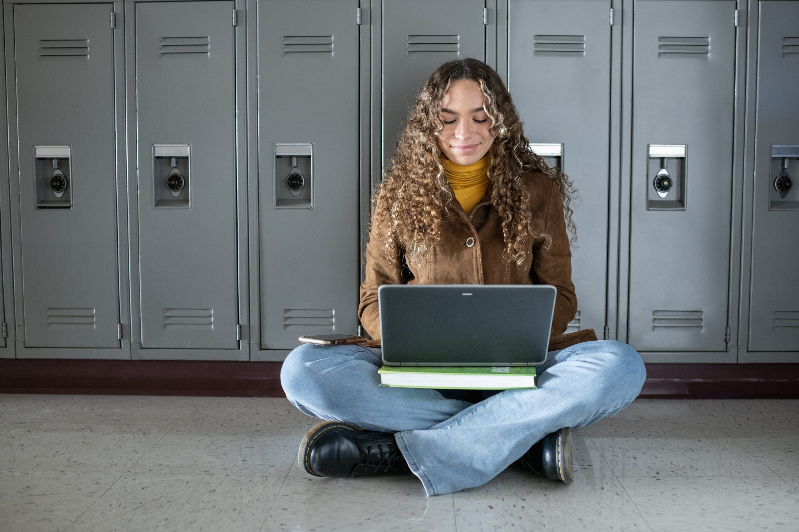 Female student sitting on floor against lockers in high school hallway using laptop