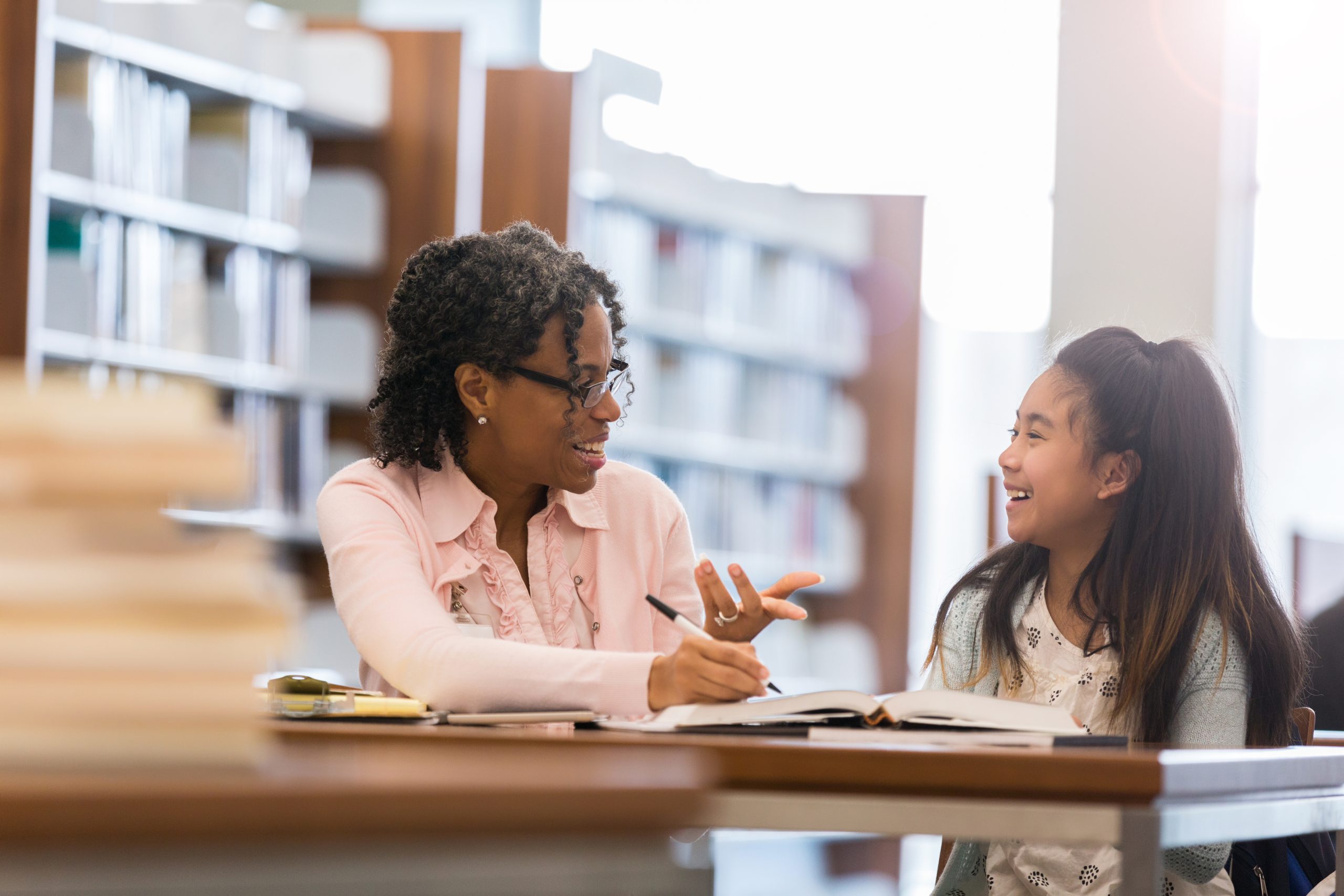 A little girl sits in her elementary school library with her mature female tutor.  She laughs as the tutor gestures and speaks.