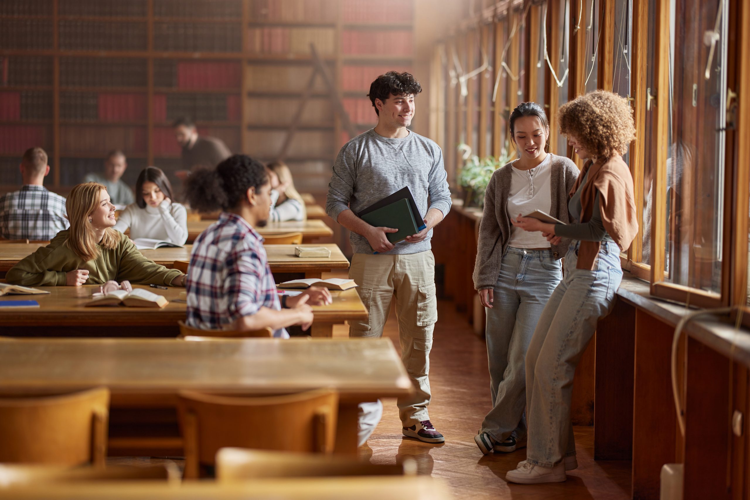 Group of happy diverse students enjoying on a break in library. One of them is using touchpad.