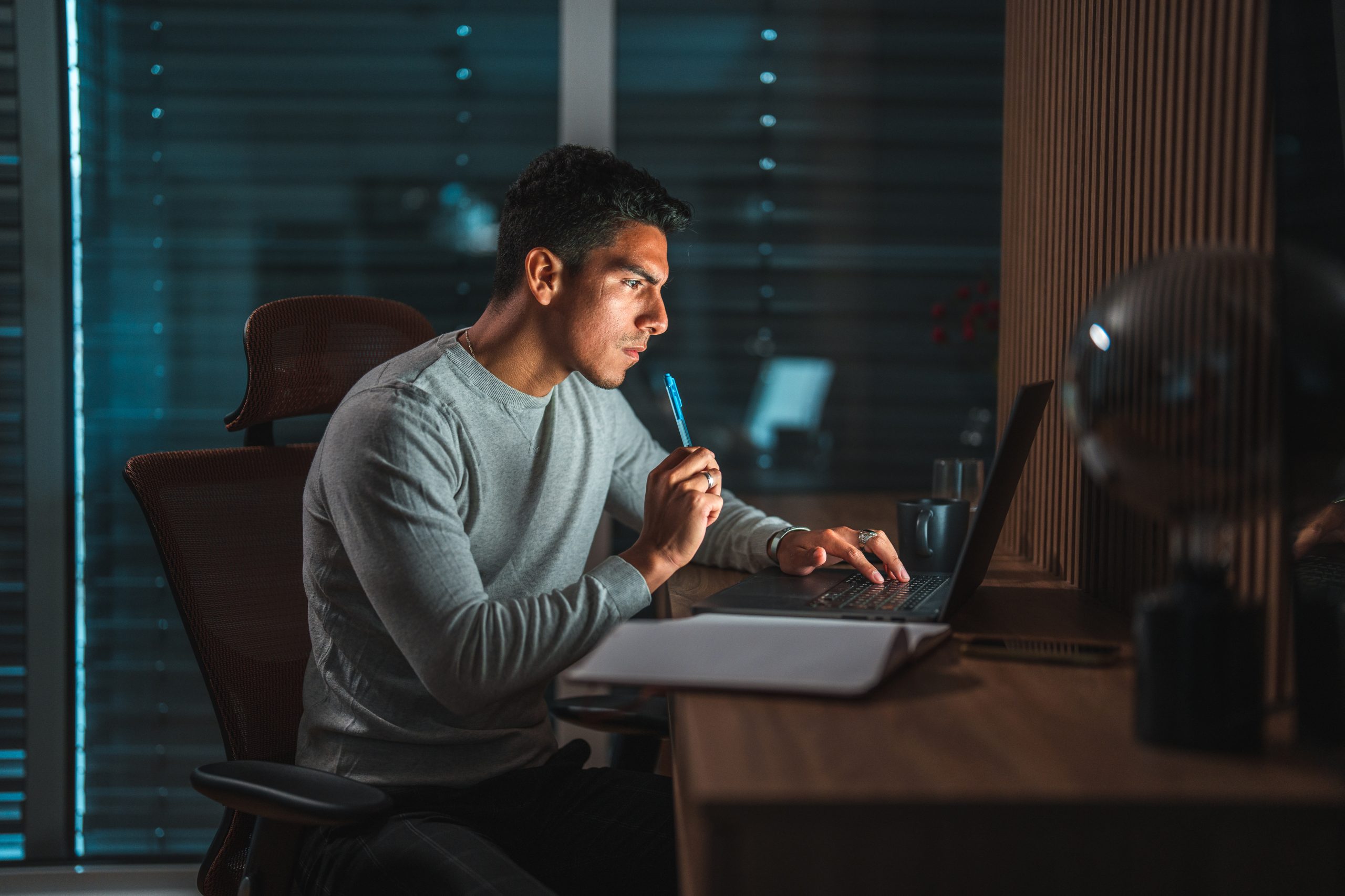 Waist up shot of a young Hispanic male entrepreneur working at night, a laptop and an open writing pad on the desk in front of him. Bright glow from the screen reflecting on his face and in the terrace window. Shutters fully closed in the background.