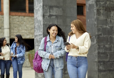 Happy young women discussing in campus. Smiling student showing smart phone to friend. They are wearing casuals.