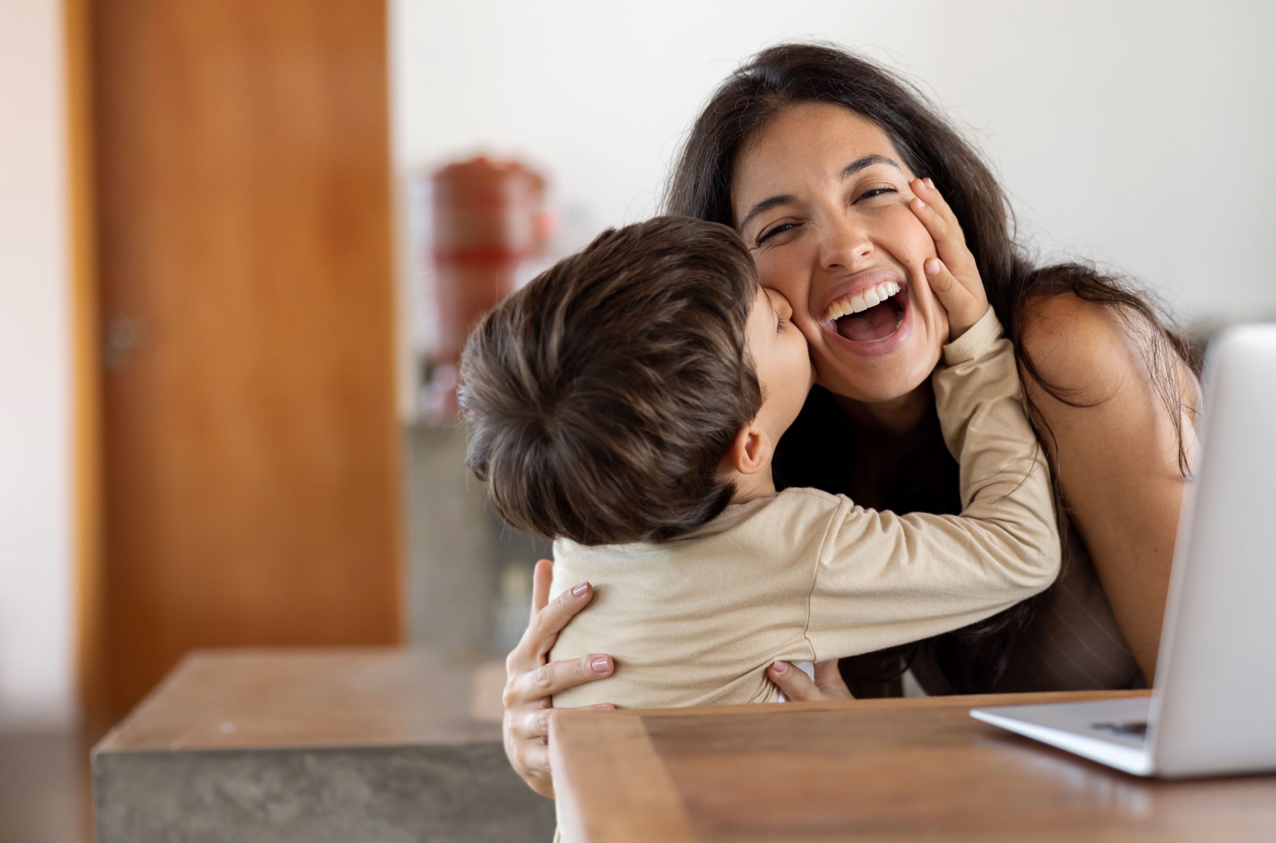 Loving son giving a kiss to her mother while she is working at home on her laptop - lifestyle concepts
