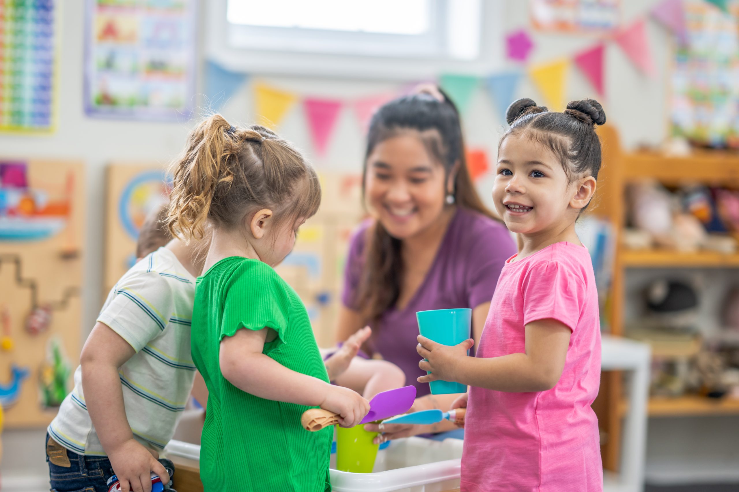 A preschool teacher knees down with a small group of children as they gather around an activity table filled with rice.  They are each dressed casually and are smiling as they enjoy playing together.