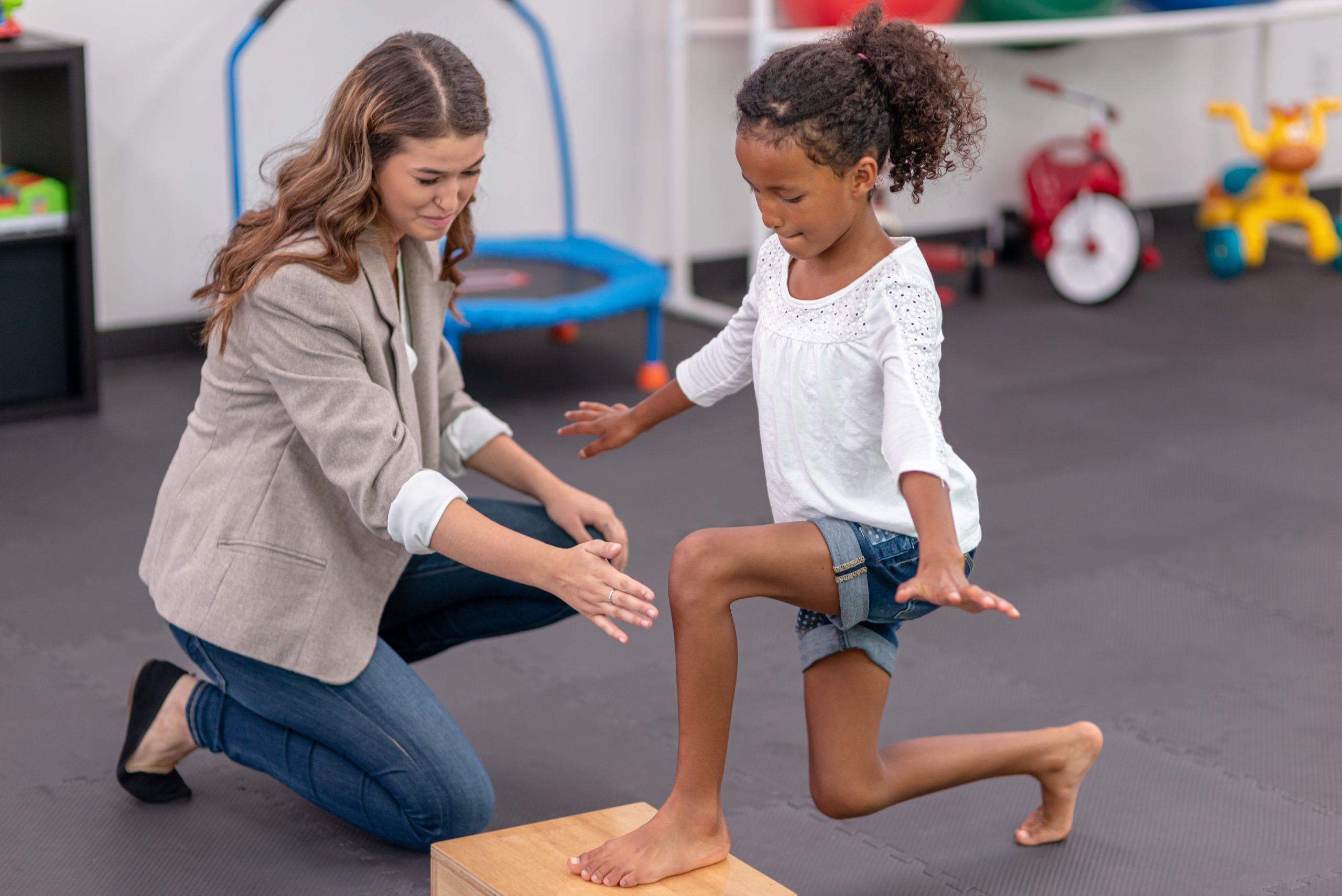 A black girl is doing lunges on a hard platform with the help of her female caucasian physical therapist. She has one foot on the block as she looks down.