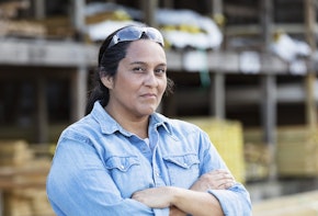 A mature Hispanic woman working in a lumberyard or building supply store, looking at the camera with her arms crossed. Shelves stacked with lumber are out of focus in the background.