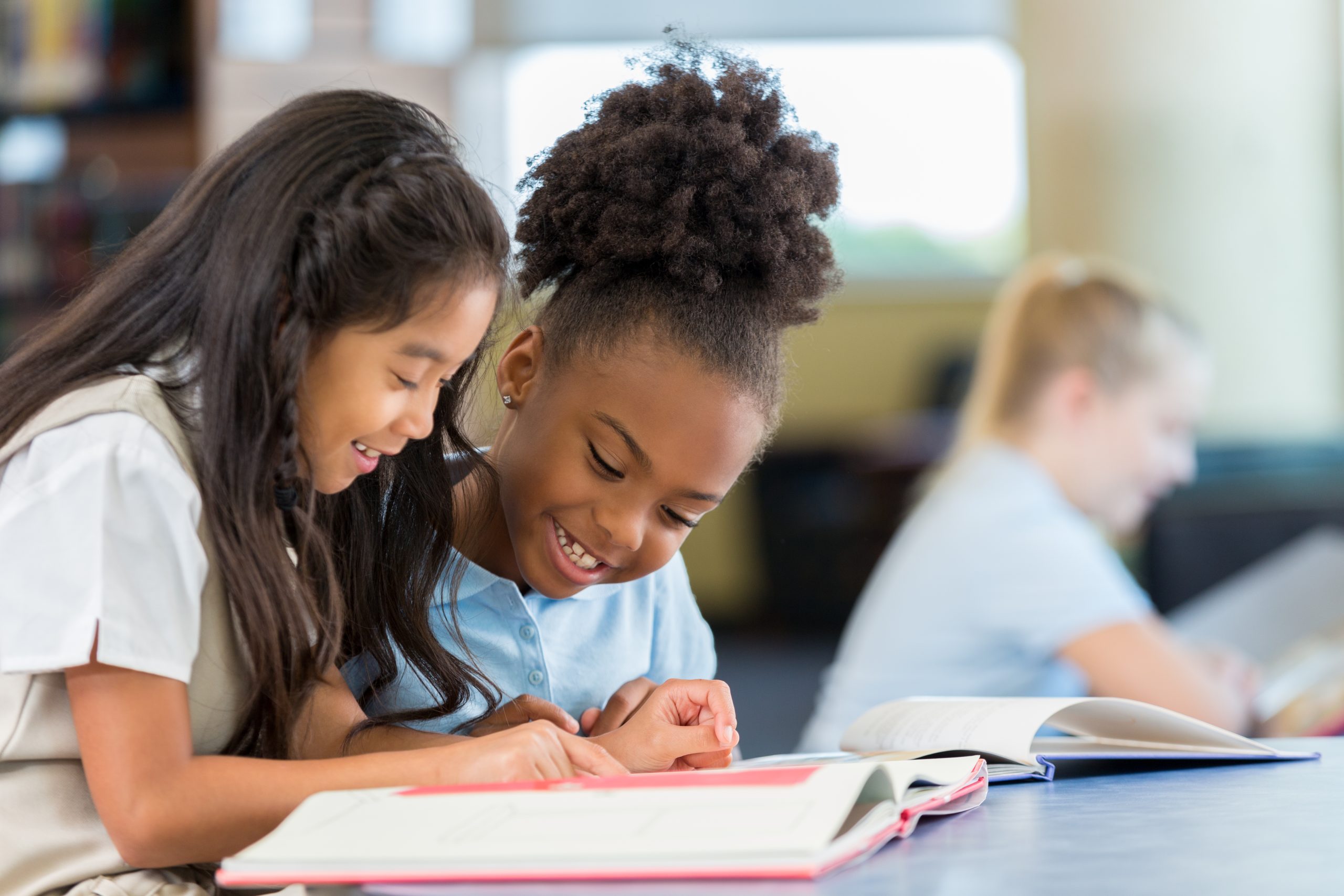 Happy female elementary age students in school uniforms are sitting at a desk in the school library and reading a book together. A Filipino elementary age girl sits with an African American classmate and reads a book together in the school library.