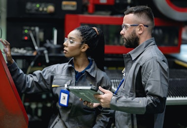 Continuous Manufacturing Improvement Can Improve Engineering Product Quality with Total Quality Management (TQM). A male industrial engineer advises a female engineer about the automatic punching machine control process for engineering parts production.